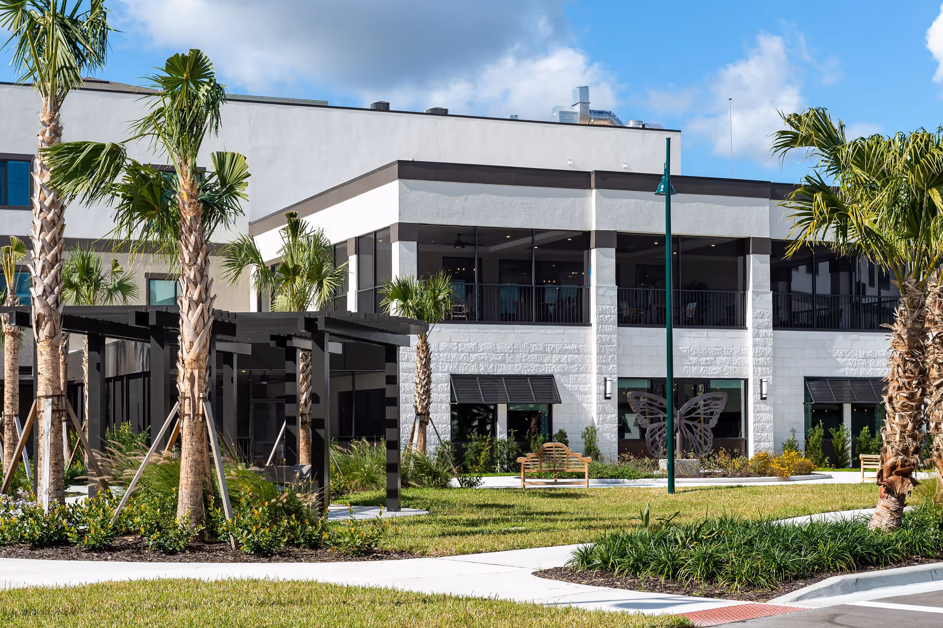 Exterior view of a modern senior living facility with palm trees, landscaped greenery, benches, and a butterfly decoration on the building's wall under a blue sky with some clouds.
