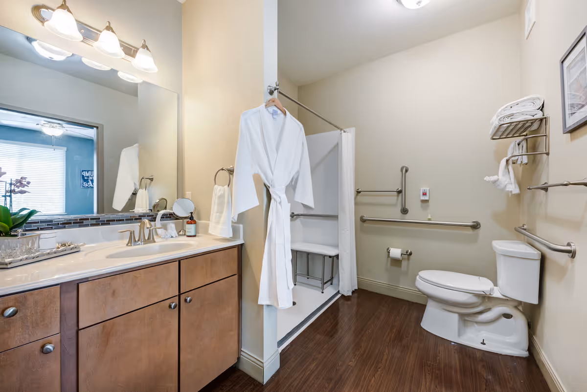 A clean and accessible bathroom with a wooden floor, a vanity with a sink and mirror, a white bathrobe hanging on a hook, a walk-in shower with a bench and grab bars, a toilet with grab bars around it, and neatly folded towels on a wall-mounted rack.