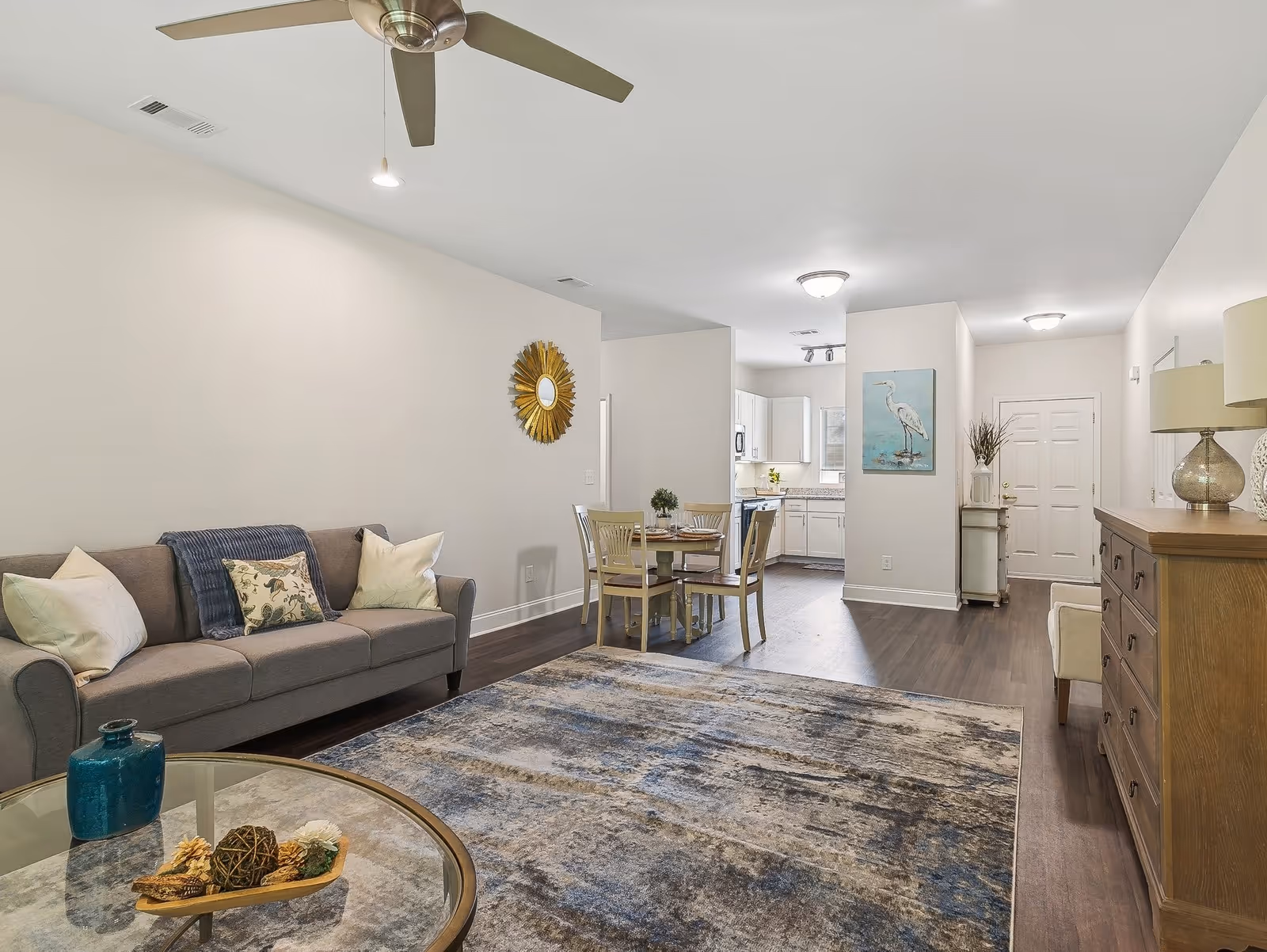 A bright and spacious living area in a senior living facility featuring a gray sofa with decorative pillows, a round glass coffee table with decorative items, a large area rug with blue and gray tones, a ceiling fan, a small dining table with four chairs, and a kitchen with white cabinets in the background. The walls are light-colored with minimal decor including a sunburst mirror and a painting of a bird.