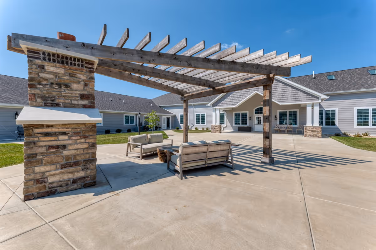 Outdoor patio area at Cedarhurst Senior Living of Highland featuring a wooden pergola with stone pillars, cushioned outdoor seating, and a concrete floor. The building exterior is visible in the background under a clear blue sky.