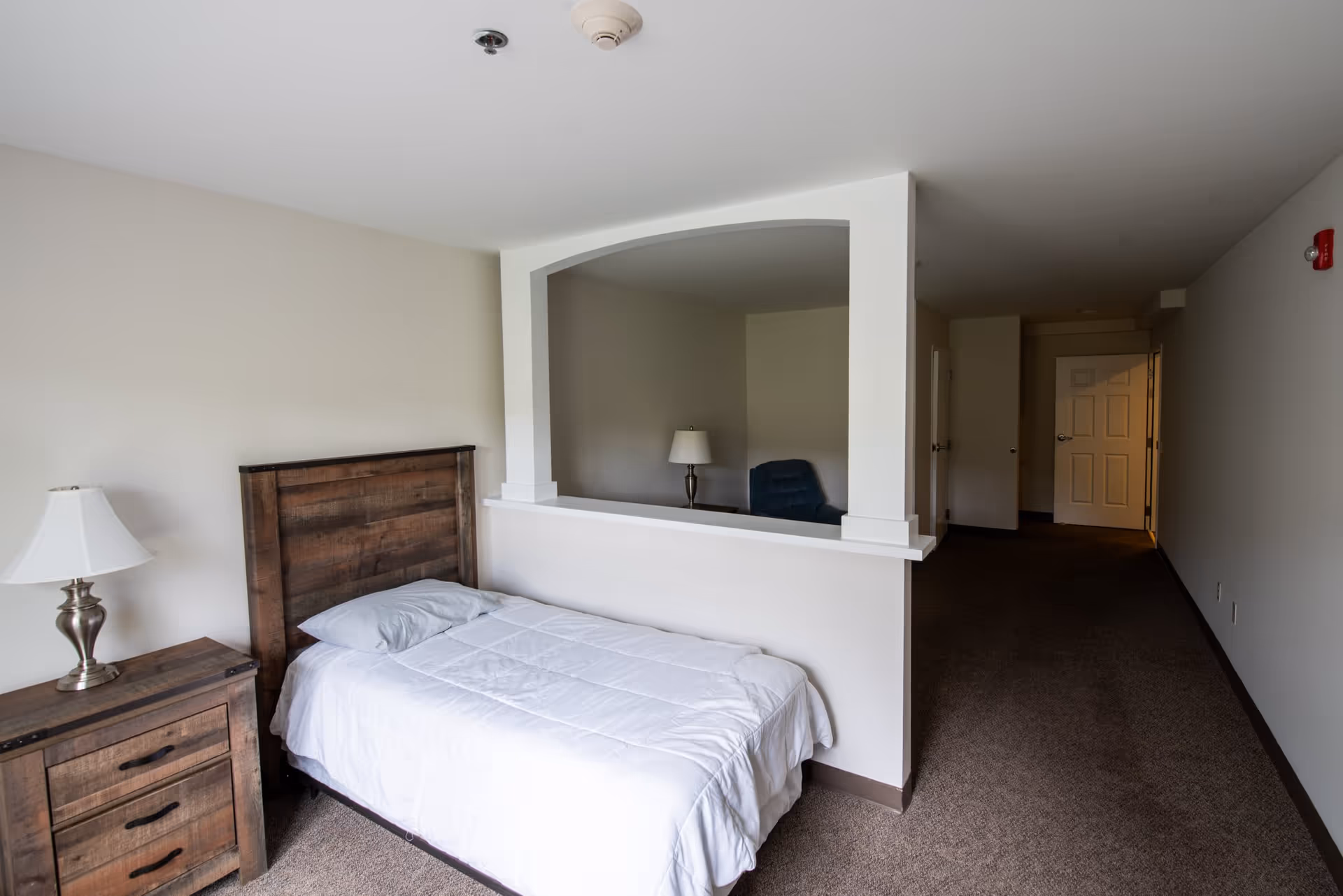 A simple single bedroom with a bed, wooden nightstand and lamp, and a half wall with an arch opening to a small sitting area and hallway.