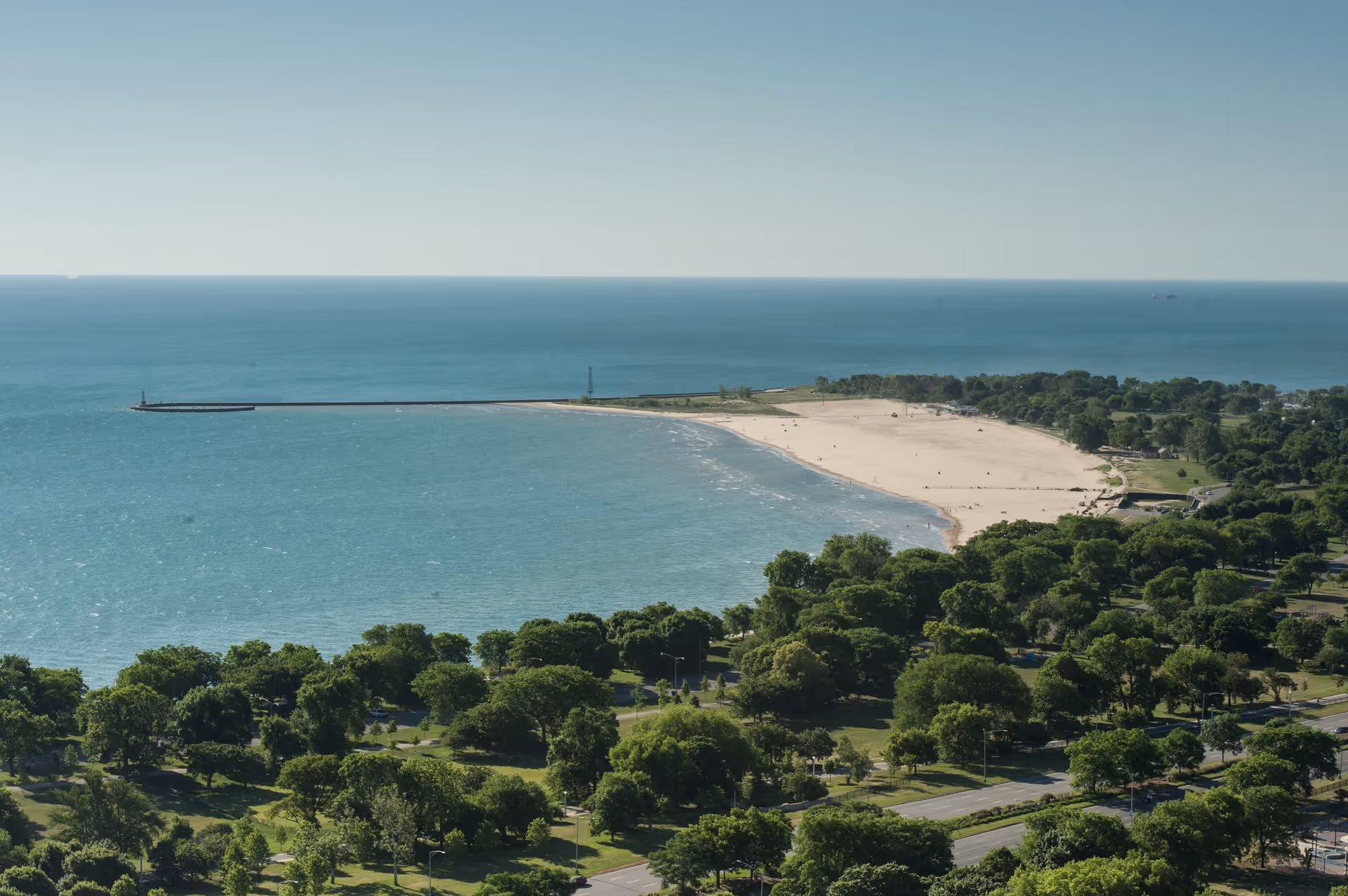 Aerial view of a large sandy beach bordered by a long pier extending into a calm blue lake, with a lush green park and trees in the foreground under a clear sky.