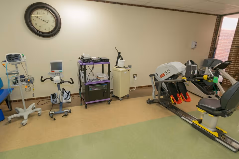A rehabilitation room with various exercise and medical equipment including a stationary bike, a SCIFIT exercise machine, medical monitors, and a clock on the wall showing 10:10. The room has beige walls, a green and tan floor, and a window showing a brick exterior.
