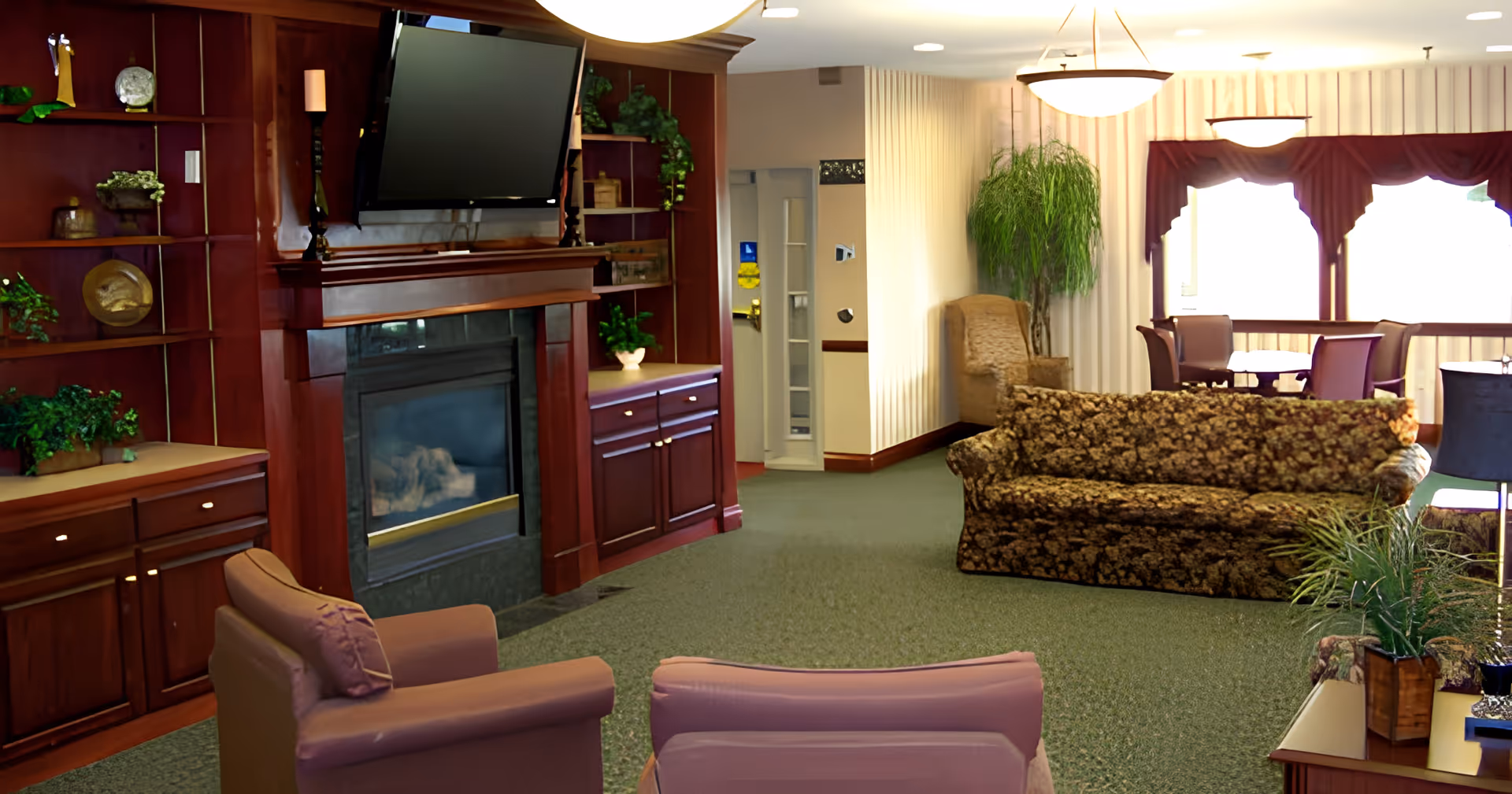 A cozy living room area in a senior living facility featuring a floral-patterned sofa, two mauve armchairs, a wooden entertainment center with a mounted flat-screen TV above a fireplace, and decorative plants. In the background, there is a dining area with tables and chairs near large windows with maroon valances, and soft overhead lighting.