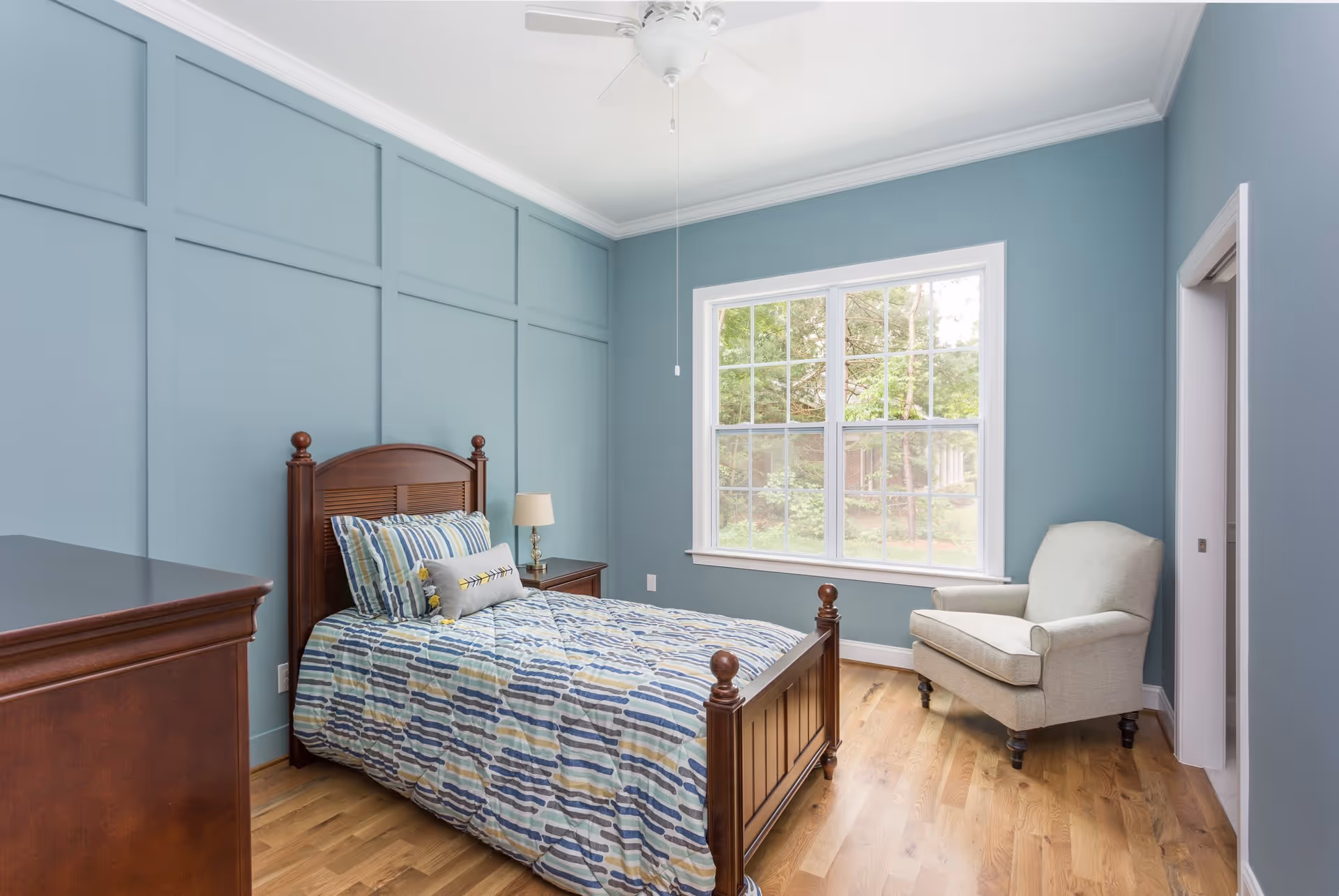 A bedroom with light blue walls and wooden floor. It features a single wooden bed with a colorful striped comforter and pillows, a wooden dresser, a nightstand with a lamp, a beige armchair near a large window showing greenery outside, and a ceiling fan above.