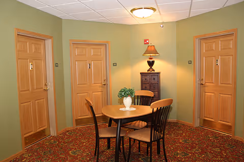 A small seating area with a round wooden table and four chairs on a patterned carpet. The walls are painted green and there are three wooden doors surrounding the area. A small wooden cabinet with a lamp and a potted plant on the table are also visible.