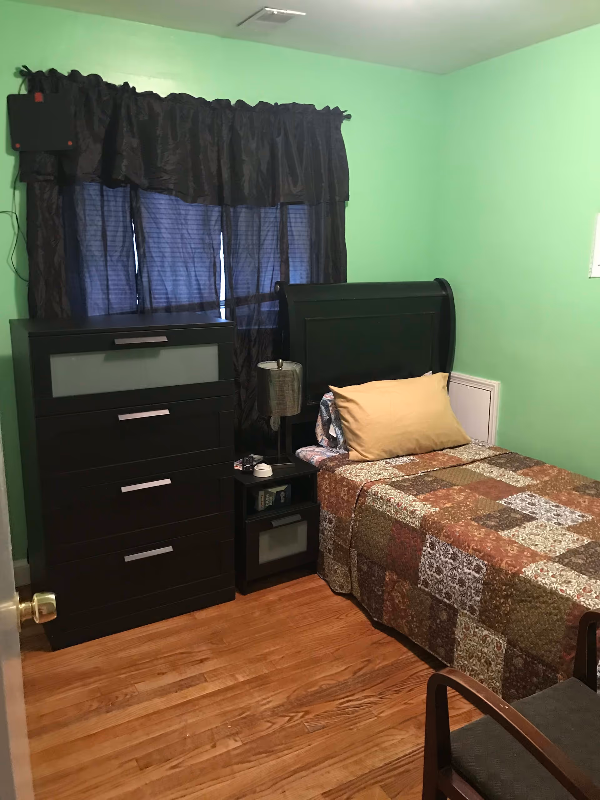 A small bedroom with light green walls and wooden floor. The room contains a single bed with a patchwork quilt and a beige pillow, a black headboard, a black nightstand with a lamp and some items on it, a black chest of drawers, and a window with black curtains.