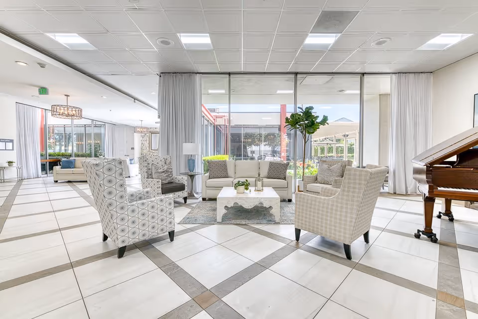 A spacious and bright living room area with large windows overlooking a garden. The room features a white sofa, four patterned armchairs arranged around a white coffee table on a rug, a floor lamp, and a grand piano on the right side. The floor is tiled with a geometric pattern, and there are white curtains framing the windows.