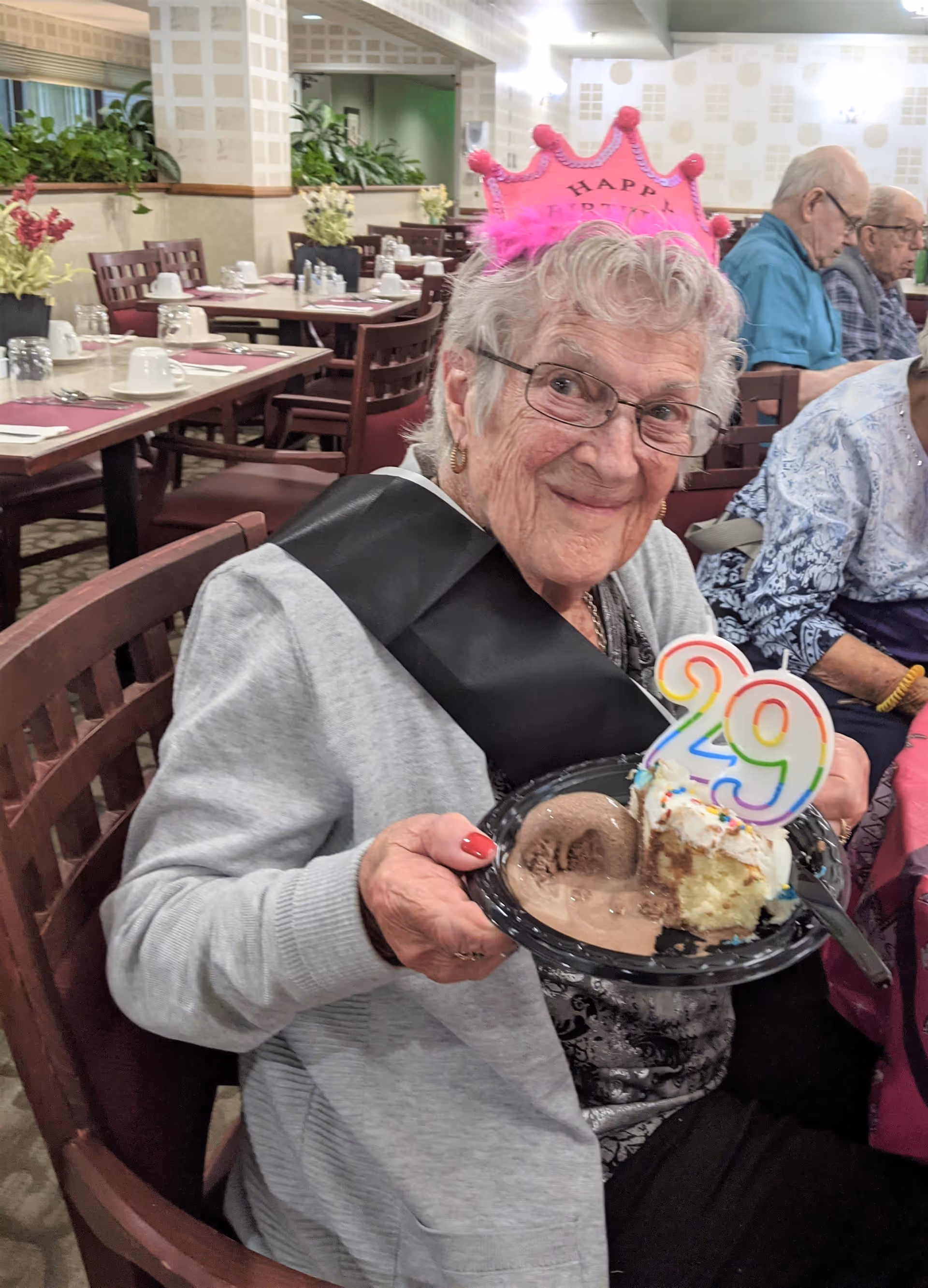 An elderly woman wearing a pink "Happy Birthday" crown holds a plate with cake and ice cream while seated in a dining room.