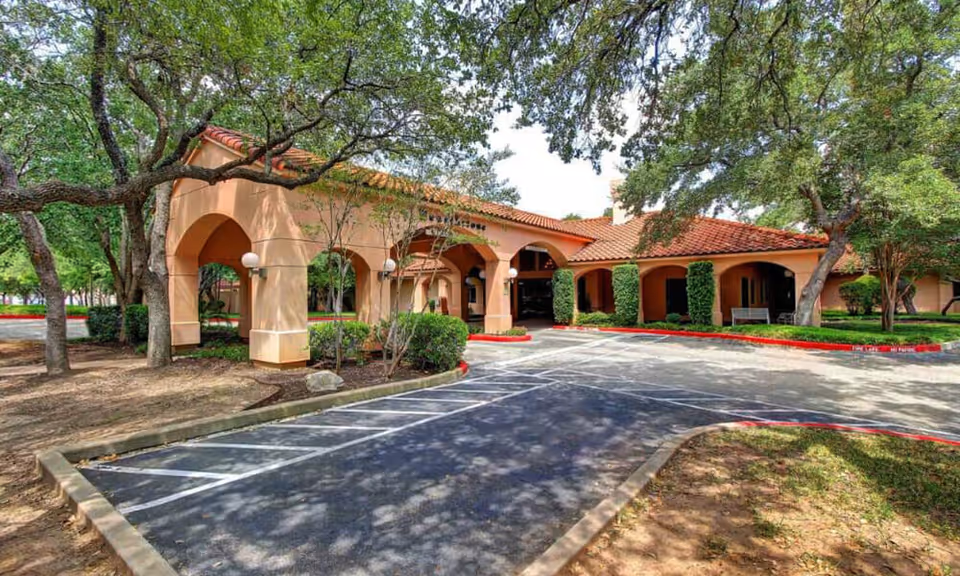 Exterior view of Hearthstone Nursing and Rehabilitation facility showing a building with a terracotta tiled roof, arched entryways, surrounded by trees and landscaping, with a driveway and parking area in front.