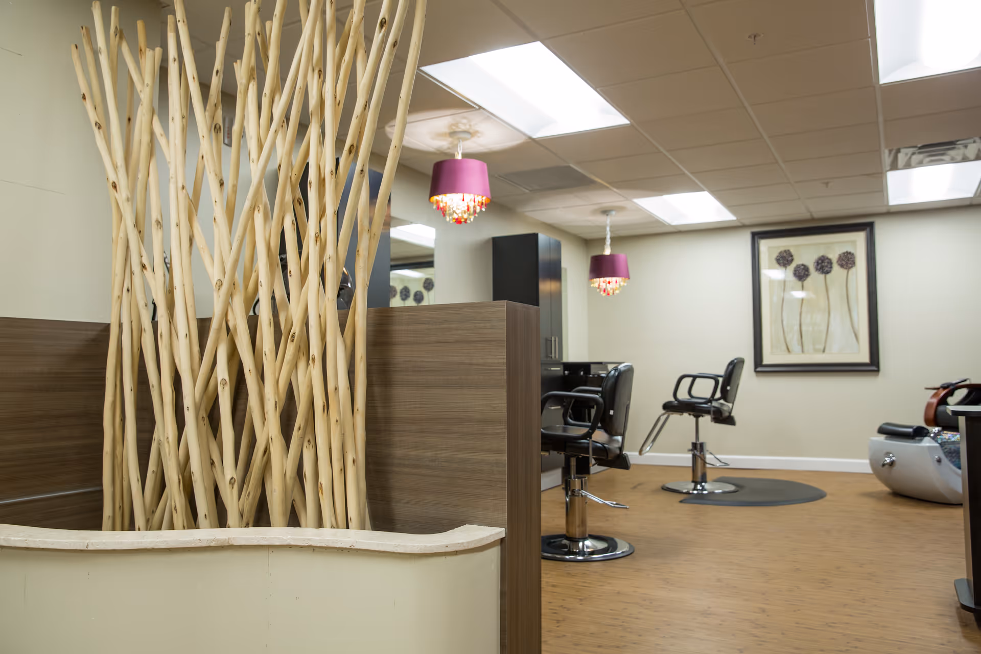 Interior salon area with styling chairs, decorative wooden stick divider, pendant lights, and framed artwork on the wall.