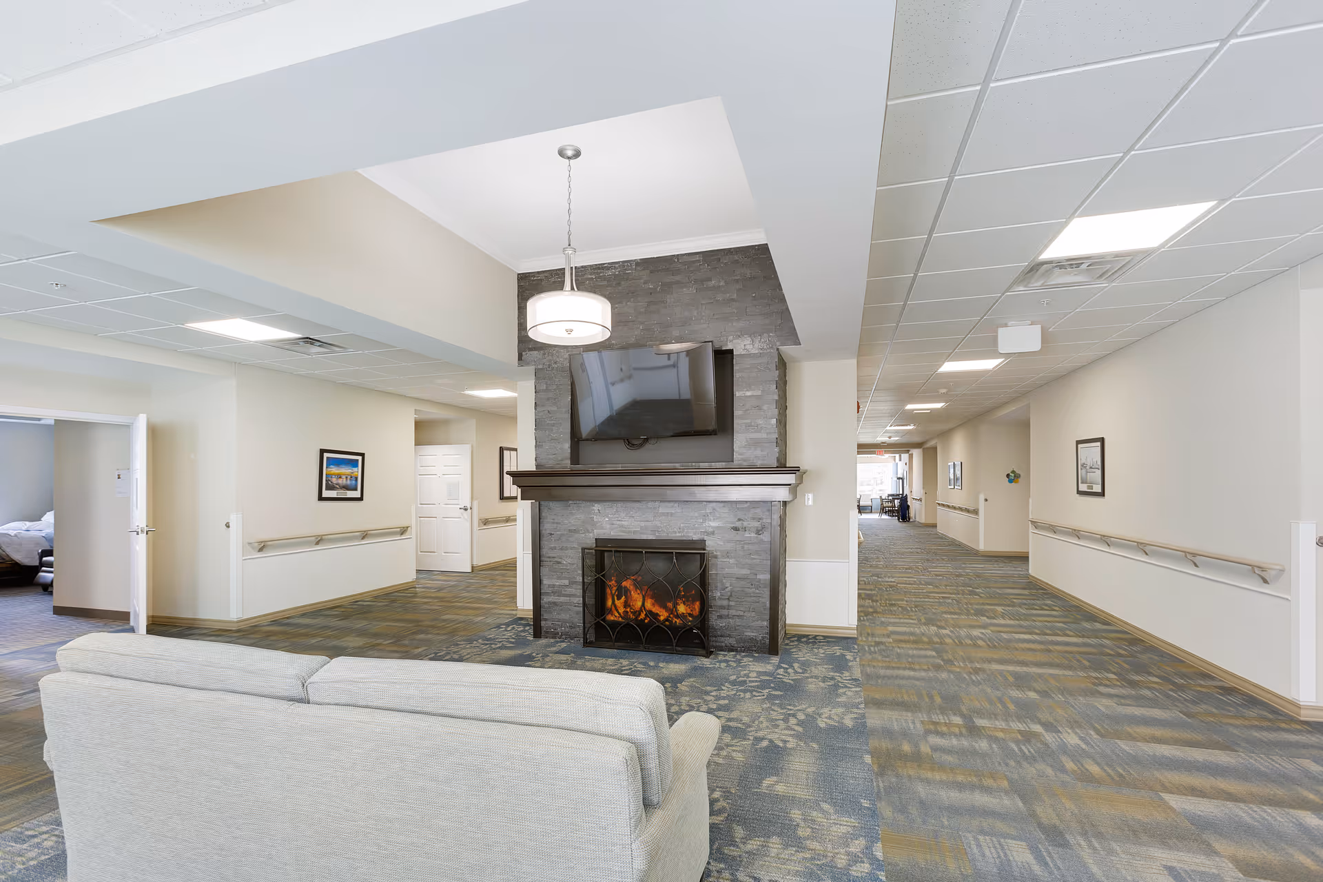 Interior view of a senior living facility hallway with a light gray couch facing a stone fireplace with a mounted flat-screen TV above it. The hallway has patterned carpet, white walls with handrails, framed pictures, and ceiling lights. Doors and additional rooms are visible along the hallway.