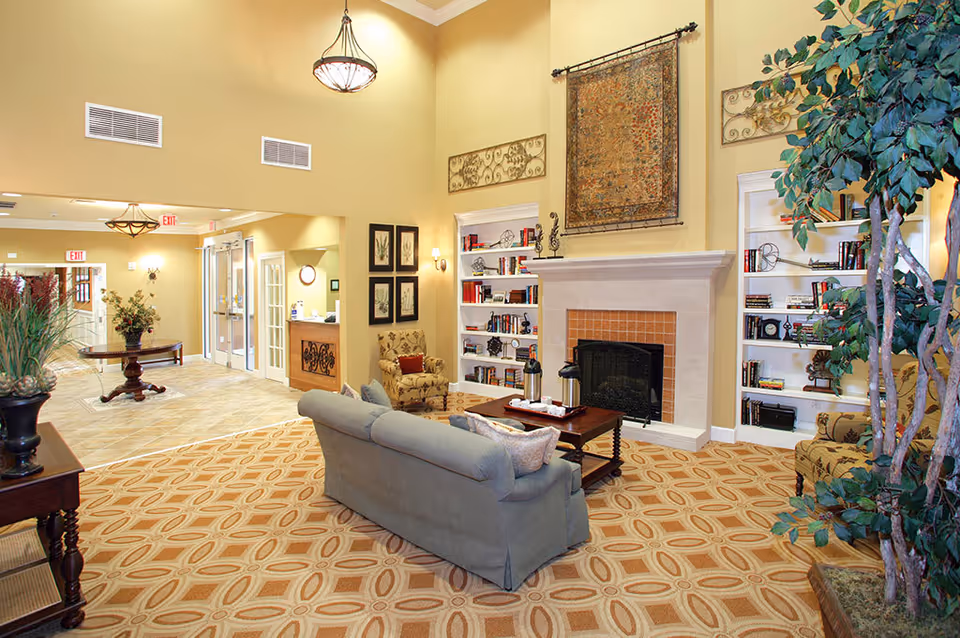 A spacious and well-lit living room area in a senior living facility featuring a beige couch, patterned armchairs, a wooden coffee table with a coffee set, built-in white bookshelves filled with books and decorative items, a fireplace with a large decorative tapestry above it, and a tall indoor plant. The room has high ceilings, warm yellow walls, and patterned carpet flooring. In the background, there is a hallway with exit signs and a reception desk.
