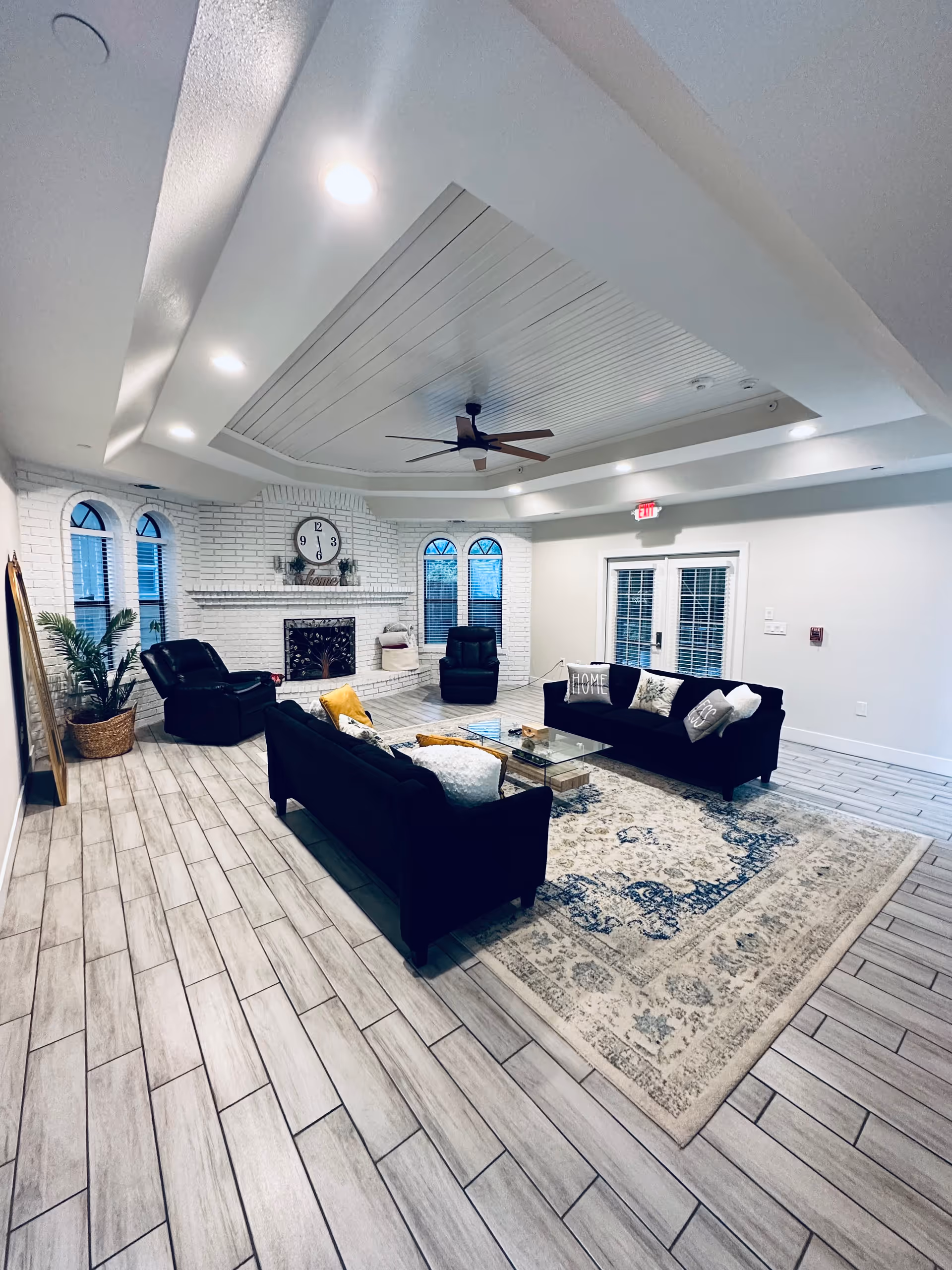 A spacious living room with light wood tile flooring and a large patterned area rug. The room features two black sofas with decorative pillows, a glass coffee table, and two black recliner chairs. There is a white brick fireplace with a clock and decorative items above it, flanked by three arched windows with blinds. The ceiling has recessed lighting and a ceiling fan. Double glass doors with blinds lead outside.