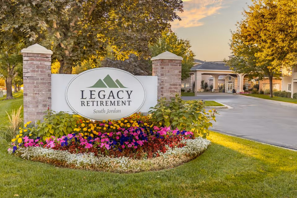 Entrance sign for Legacy Retirement South Jordan surrounded by colorful flowers and greenery, with a driveway and building visible in the background under a partly cloudy sky.