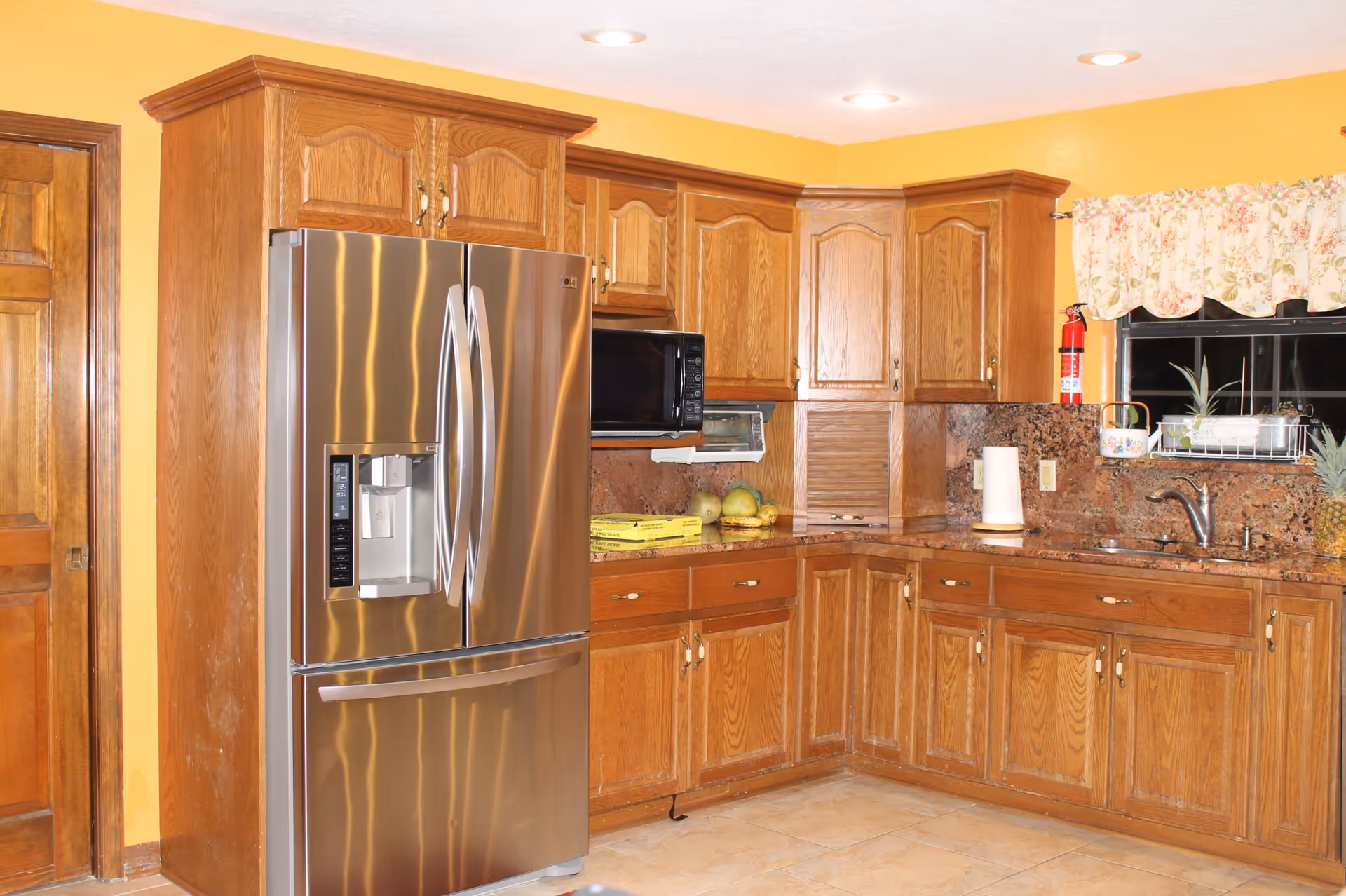 A kitchen with wooden cabinets and drawers, a stainless steel refrigerator with a water and ice dispenser, a microwave oven, a toaster oven, a granite countertop, a sink under a window with floral curtains, a fire extinguisher mounted on the wall, and some fruits and paper towels on the counter.