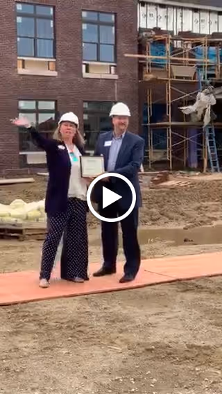 Two people wearing white construction helmets stand on a wooden plank in front of a building under construction. One person is holding a certificate and the other is gesturing with their hand. The building behind them has brick walls and large windows, with scaffolding and construction materials visible.
