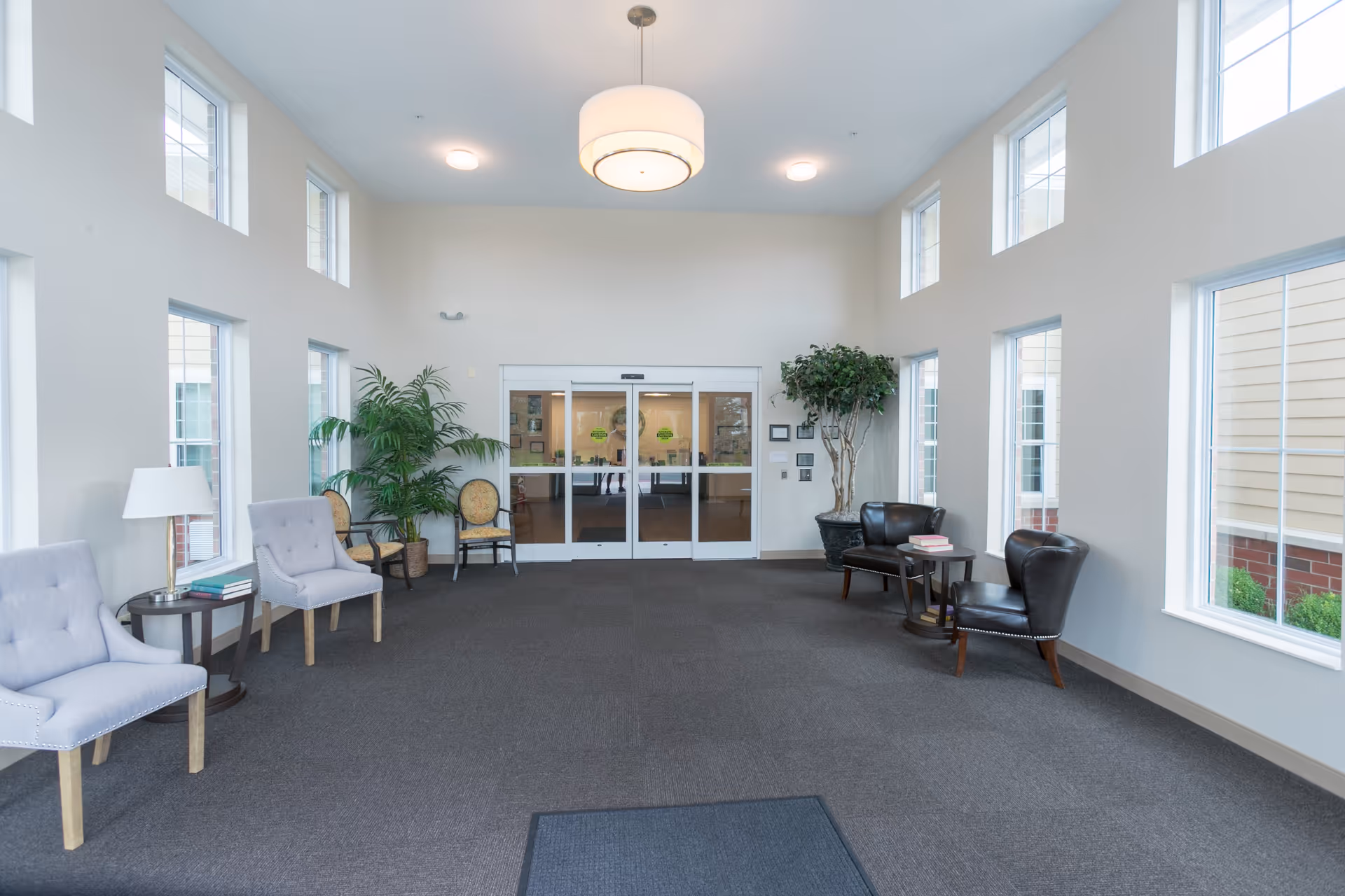 A bright and spacious waiting area with large windows on three walls allowing natural light to fill the room. The room features a dark carpeted floor, light-colored walls, and a modern ceiling light fixture. There are several chairs arranged along the walls, including two gray upholstered chairs with a small round table and lamp between them on the left, and two black leather chairs with a small table and books on the right. Two large potted plants are placed near the double glass doors at the far end of the room.