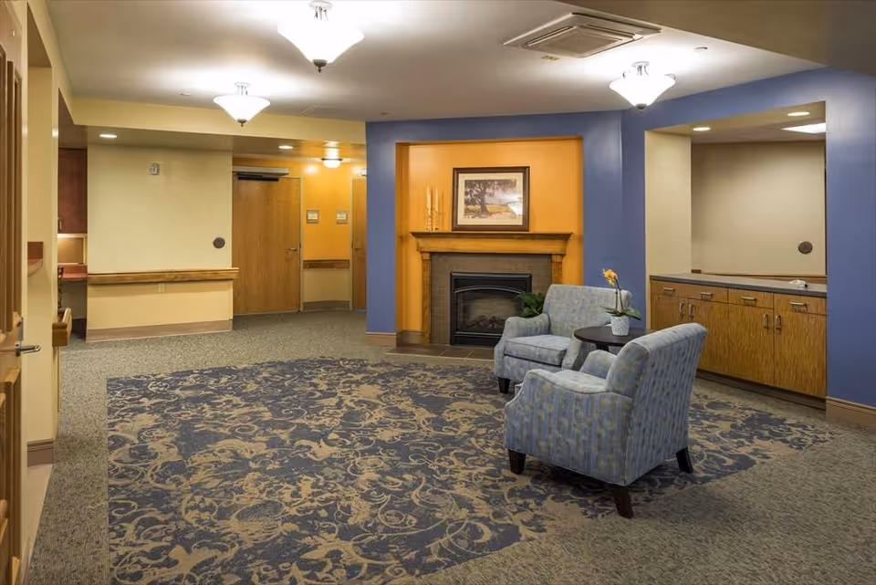 A cozy interior common area with two upholstered armchairs and a small round table with a potted plant between them. The room features a decorative fireplace with a framed picture above it, warm yellow and blue walls, patterned carpet, wooden cabinetry, and ceiling light fixtures.