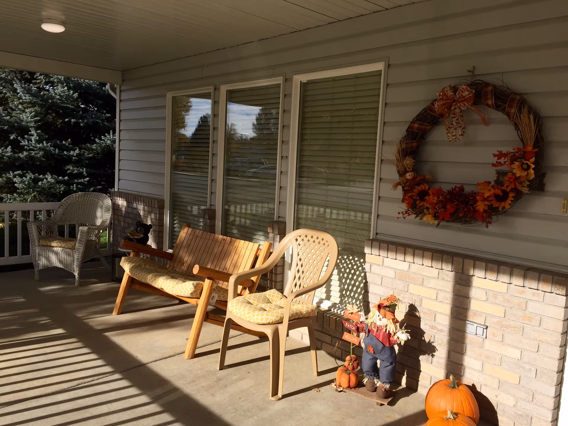 Sunlit covered front porch with chairs, a fall wreath on the wall, and pumpkins and seasonal decorations on the floor.