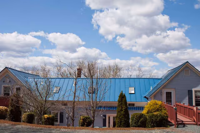 Light-blue building with a bright blue metal roof, entrance steps, shrubs and bare trees under a partly cloudy sky.
