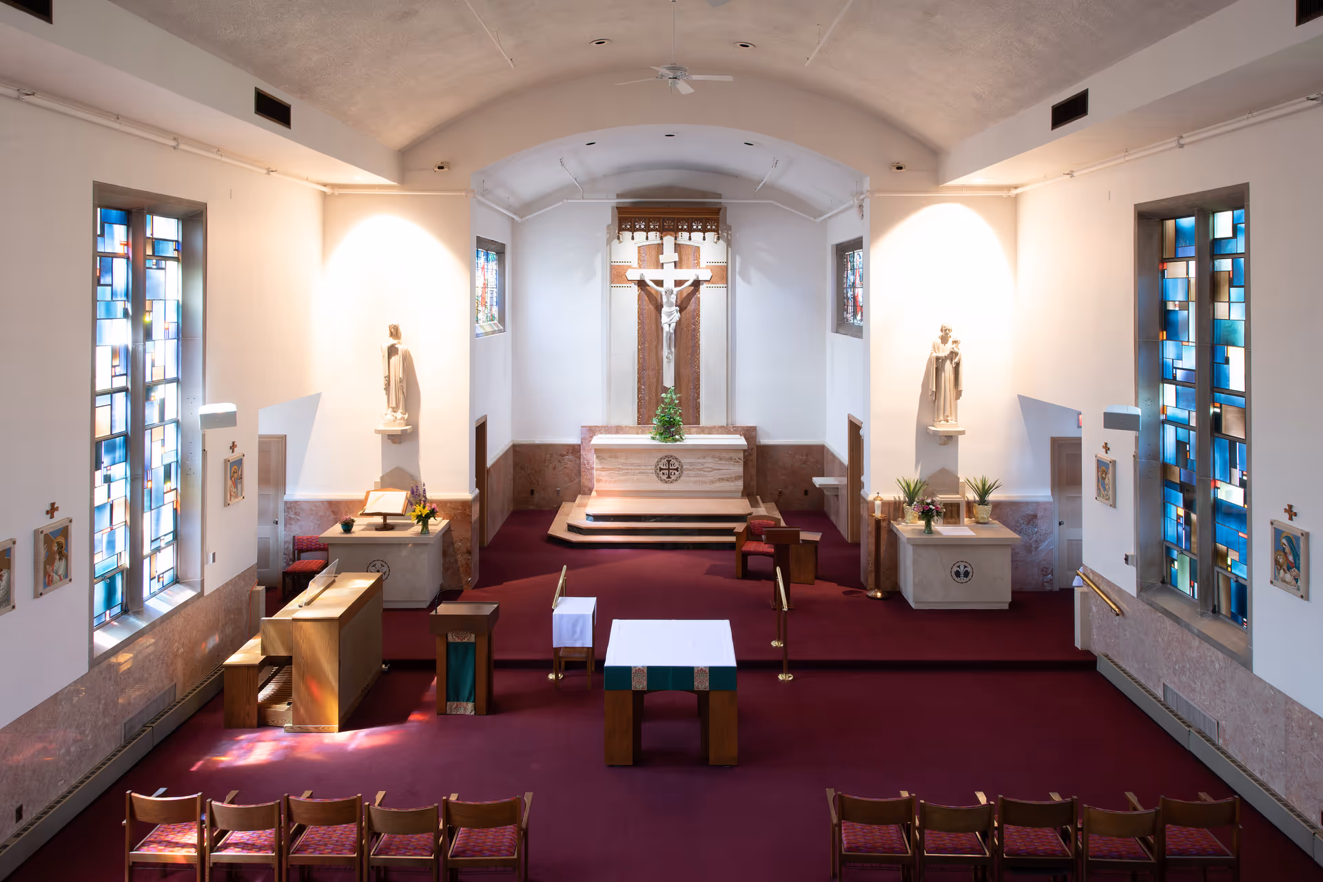 Interior view of a chapel showing an altar with a crucifix, stained glass windows, and rows of chairs.