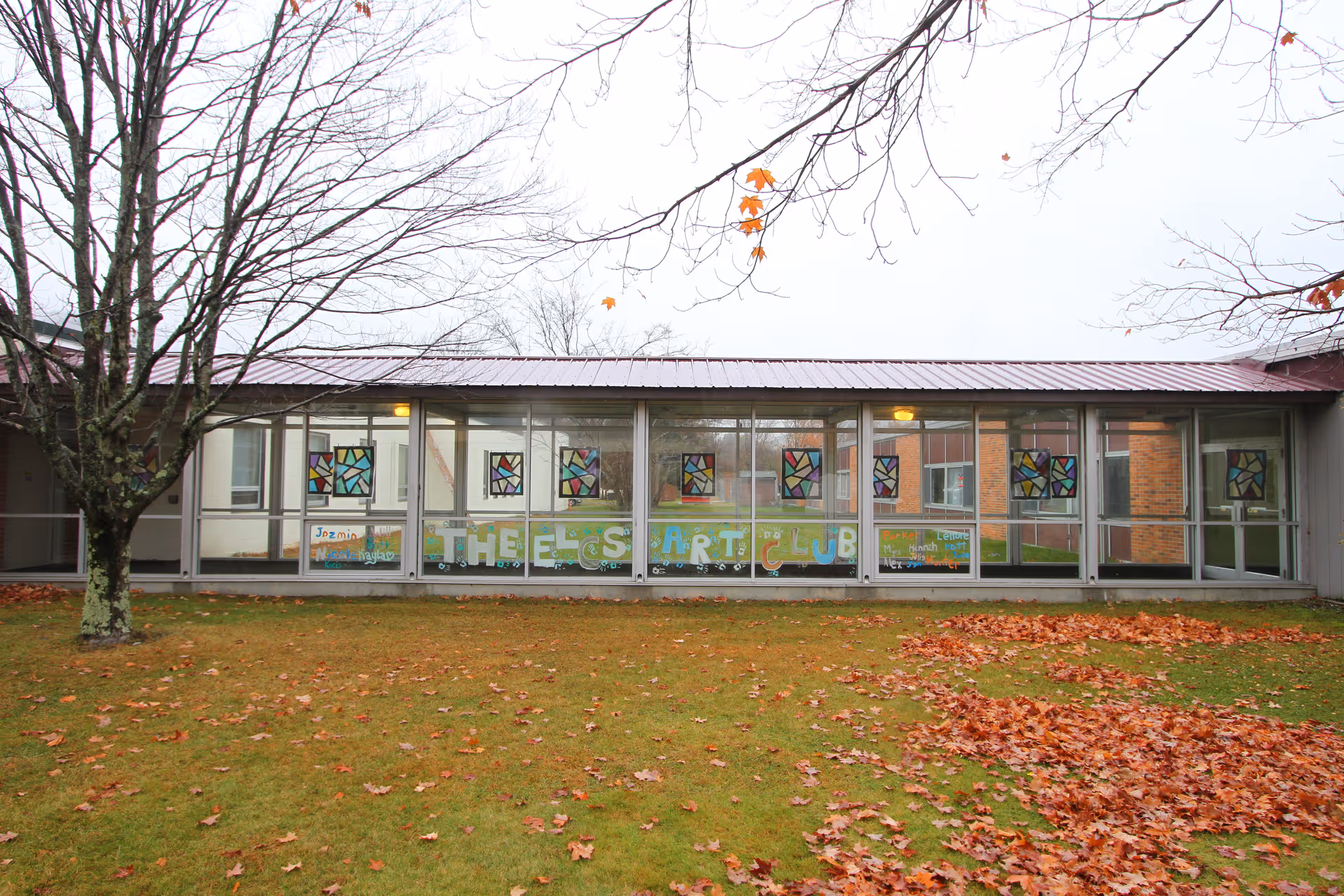 Exterior view of a single-story building with large glass windows decorated with colorful art, a grassy lawn and fallen leaves and bare trees in front.