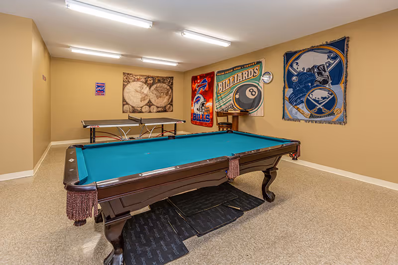 Recreation room with a green felt pool table in the foreground and a ping pong table in the background. The walls are decorated with sports-themed banners including a Bills football banner, a billiards banner, and a hockey banner. The room has beige walls and a beige floor.