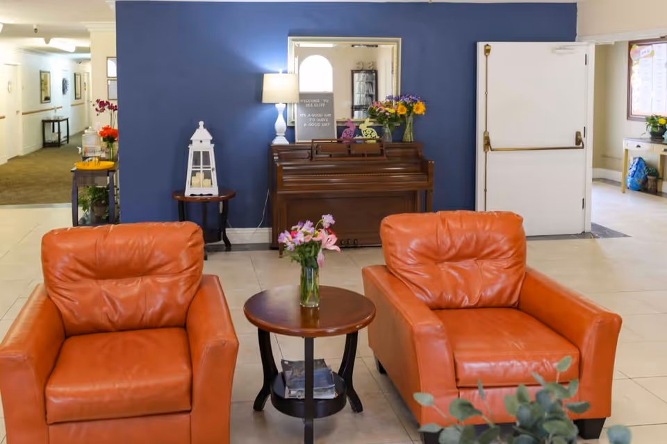 A seating area with two orange leather armchairs flanking a round table with a vase of flowers, set in front of a piano and blue accent wall in an assisted living facility lobby.