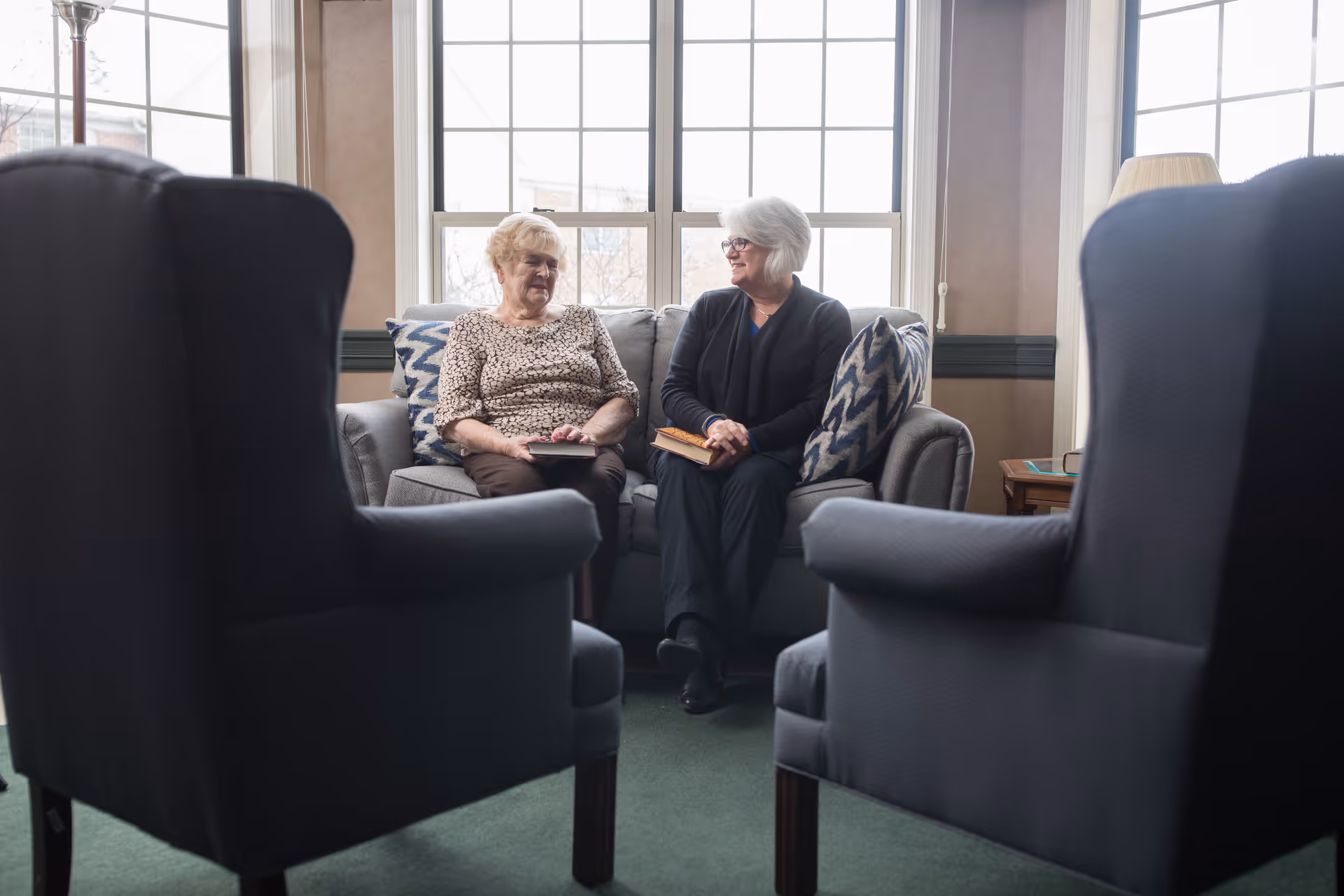 Two older women sit and talk on a sofa in a bright communal living room with armchairs in the foreground.
