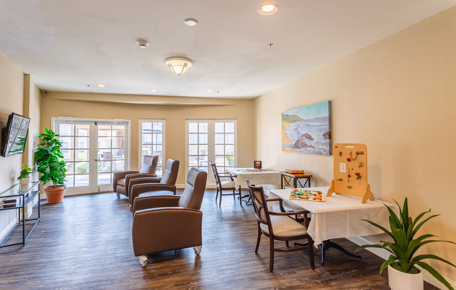 A bright and spacious common area in a senior living facility with three brown armchairs facing a wall-mounted TV, two tables with chairs, a puzzle and a game on the tables, potted plants, and large windows and glass doors letting in natural light.