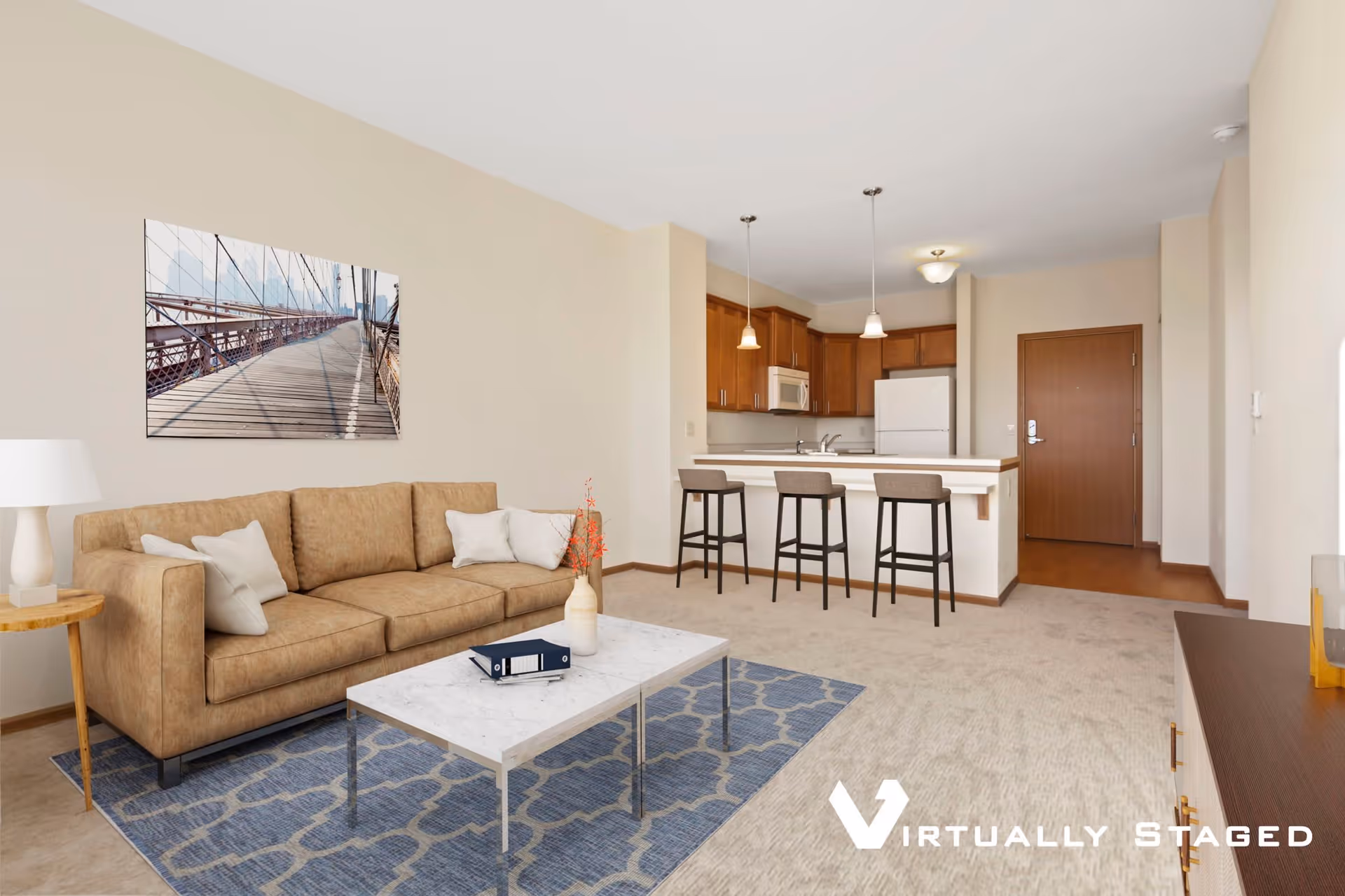 A virtually staged living room and kitchen area in a senior living facility. The living room features a tan sofa with white pillows, a white marble coffee table on a blue patterned rug, and a side table with a white lamp. On the wall above the sofa is a large framed photo of a bridge. The kitchen has wooden cabinets, a white refrigerator, microwave, and a counter with three bar stools. The entrance door is visible in the background.