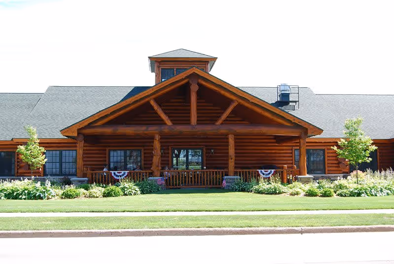 Front exterior view of a large log cabin style building with a peaked roof and a covered porch. The porch has wooden railings and is decorated with patriotic bunting and small American flags. There are well-maintained green lawns and landscaped bushes and small trees in front of the building.