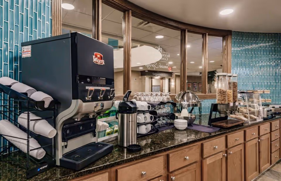 A self-serve breakfast station with a coffee dispenser, cups, cereal dispensers and pastries on a granite countertop against a teal tiled wall and mirrored windows.