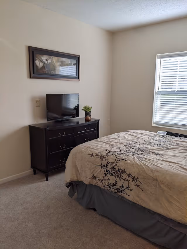 A bedroom with a bed covered in a beige comforter with a black floral pattern. Next to the bed is a dark wooden dresser with six drawers, on top of which sits a small flat-screen TV and a small potted plant. Above the dresser hangs a framed floral painting. A window with white blinds allows natural light into the room.