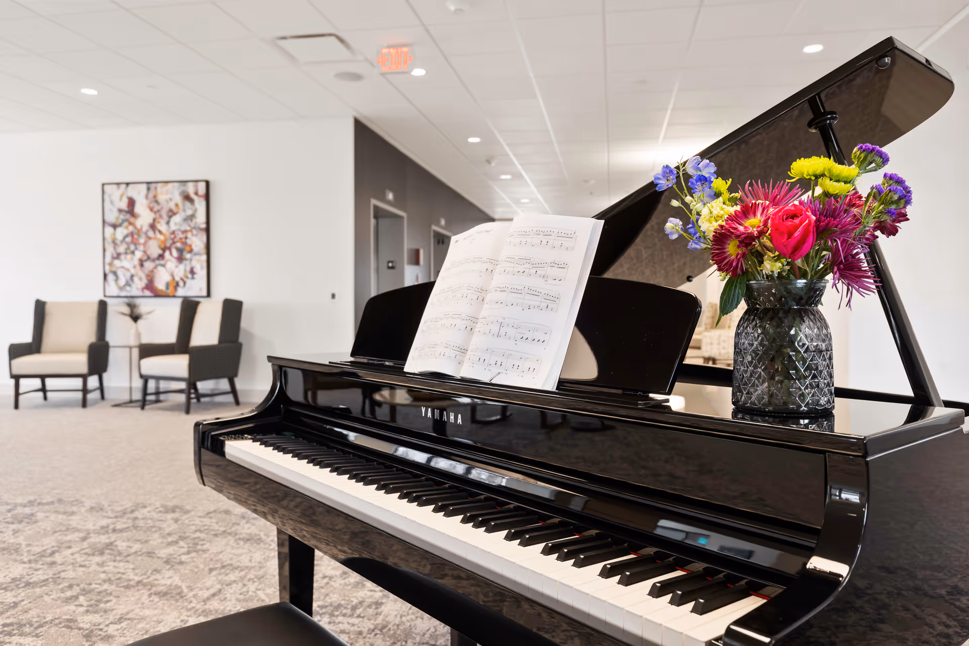 A black Yamaha grand piano with sheet music and a vase of colorful flowers on top, situated in a bright, modern common area with two armchairs and a small table in the background, along with a colorful abstract painting on the wall.