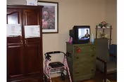 A room with a wooden wardrobe, a framed floral picture on the wall, a pink walker with a black seat, a green wooden dresser with a television on top, and a chair next to the dresser. There is also a small shelf with decorative items in the corner.