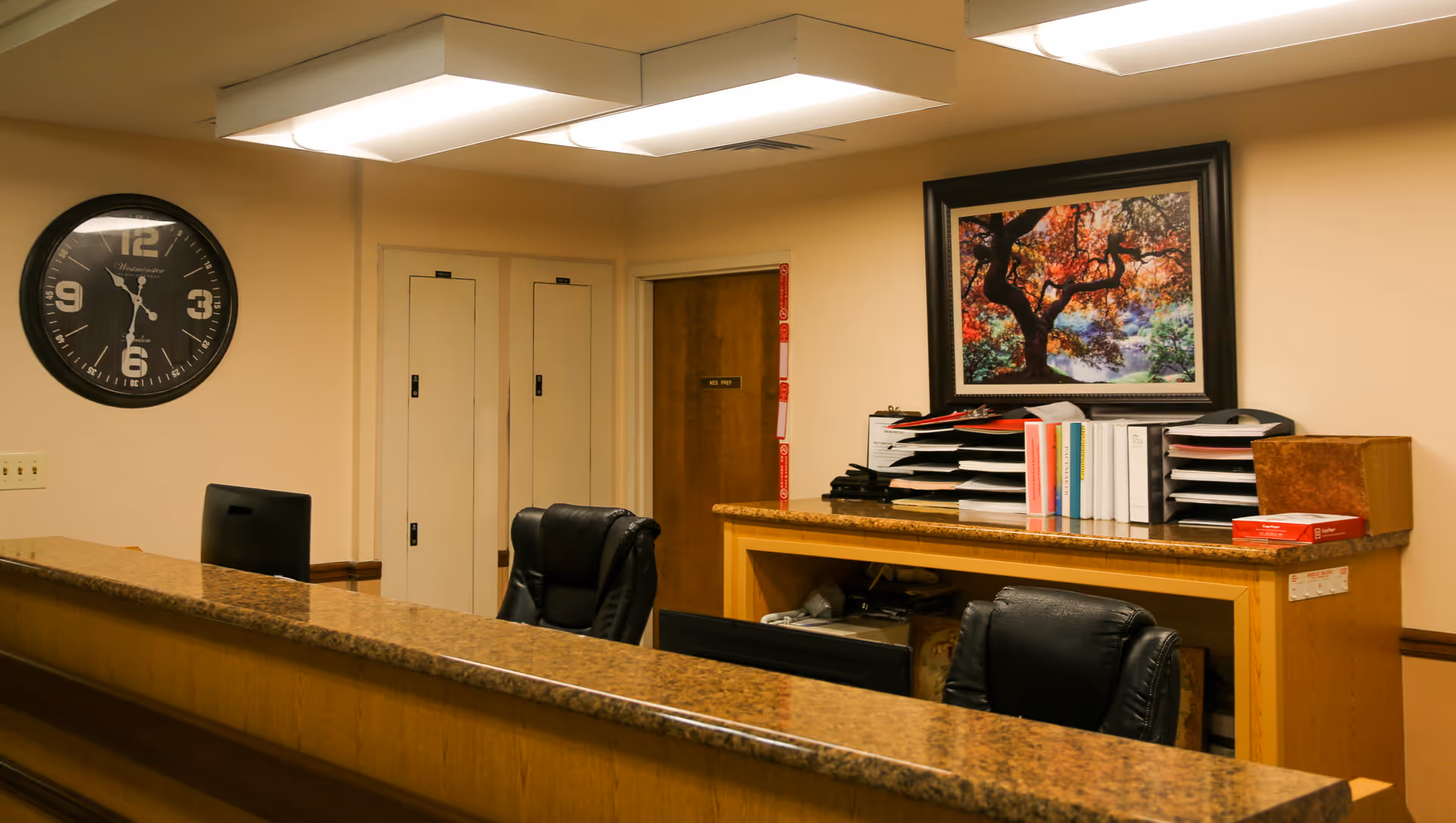 Reception area with a granite countertop desk, two black office chairs, a wall clock showing 6:15, a wooden door labeled 'Men's Restroom', file organizers with documents and binders, and a framed picture of a tree with autumn leaves on the wall.