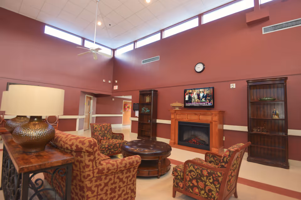 A cozy living room area with patterned armchairs and a sofa arranged around a round ottoman. There is a wooden fireplace with a TV mounted above it, two wooden bookshelves on either side, and a ceiling fan hanging from the high ceiling with windows near the top of the walls. The walls are painted a warm reddish color.