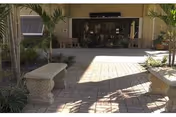 Outdoor patio area with stone benches, potted plants, and a paved brick floor leading to the entrance of a building with glass doors and windows.