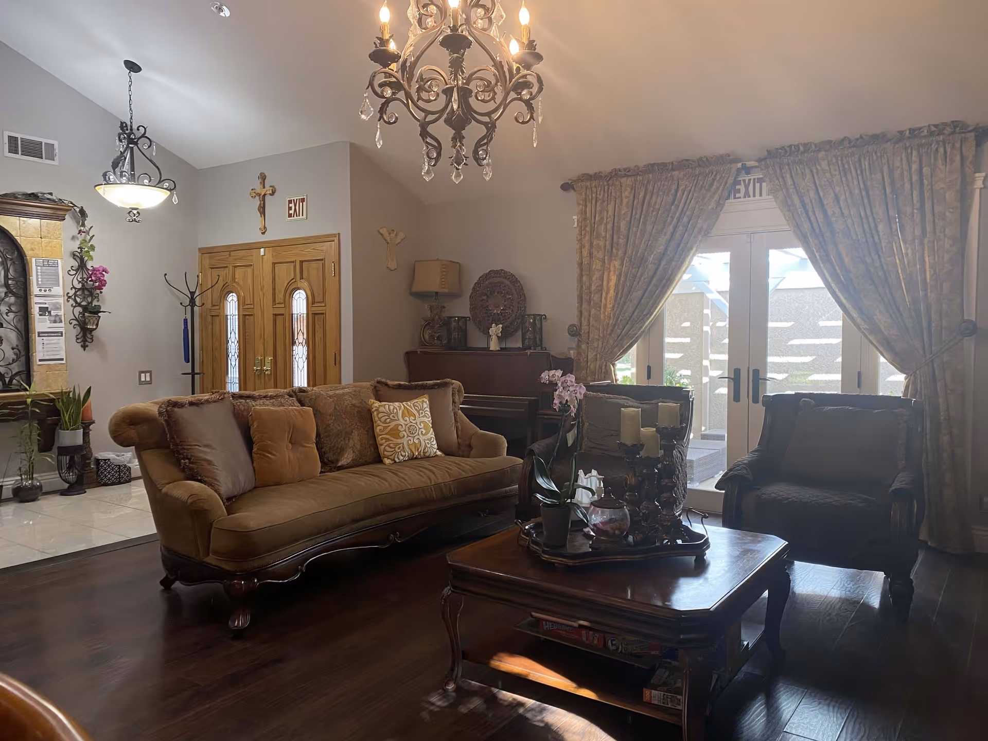 A cozy living room with a brown upholstered sofa and two armchairs around a wooden coffee table. The coffee table has decorative items including candles and a potted orchid. Behind the sofa is a wooden piano with a lamp and decorative pieces on top. The room has dark wooden flooring, beige walls, and large windows covered with patterned curtains letting in natural light. There are two exit signs visible above doors, and a chandelier hangs from the ceiling.