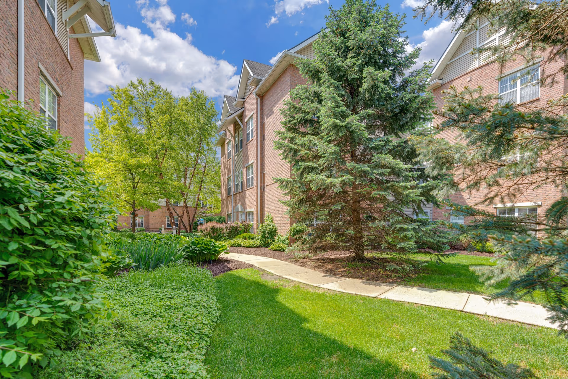 A landscaped outdoor area between brick buildings with a concrete walkway, green grass, various trees, and shrubs under a partly cloudy blue sky.