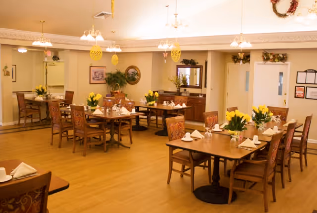 A well-lit dining room with several wooden tables and chairs arranged neatly. Each table is set with white napkins, cups, and small vases of yellow flowers. The room has light-colored walls, decorative wall hangings, and ceiling lights. There is a sideboard with a mirror and some greenery, and a menu is visible on the wall near a door.