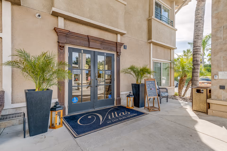 Entrance to Villas at San Bernardino facility with double glass doors framed by decorative wood trim, two large potted plants on either side, a welcome mat with the facility name, a small chalkboard sign, and outdoor seating under a covered walkway.