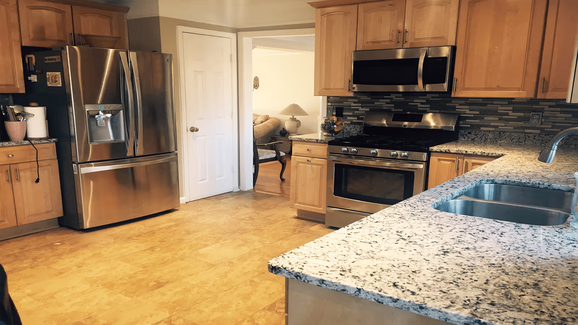 A modern kitchen with light wood cabinets, granite countertops, a stainless steel refrigerator, stove, microwave, and a double sink. The kitchen floor is a light brown tile, and there is an open doorway leading to a living room area with a beige sofa and a lamp.