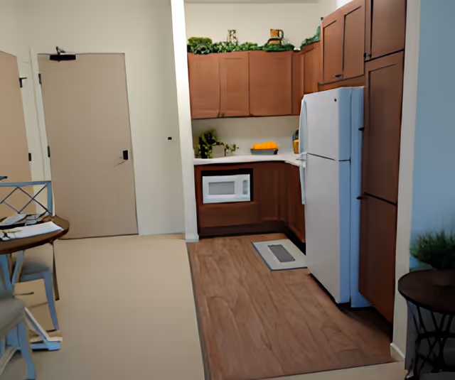 Interior view of a small kitchen area with wooden cabinets, a white refrigerator, a microwave, and some decorative plants on top of the cabinets. To the left, there is a door and part of a dining table with chairs.