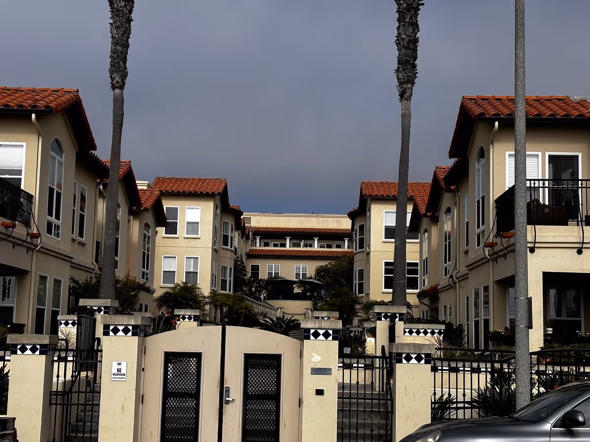 Gated courtyard entrance of a Mediterranean-style multi-story senior living complex with palm trees and red tile roofs under a cloudy sky.