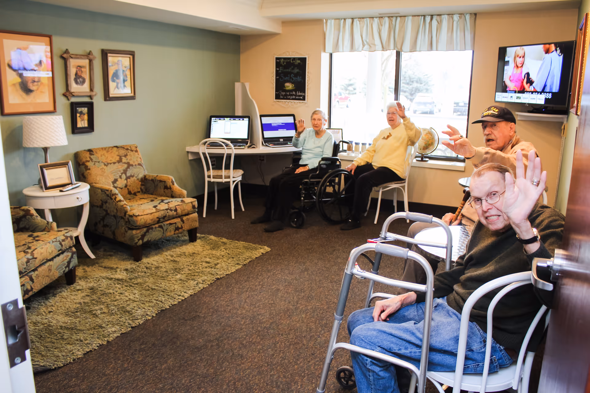 A cozy common room in an assisted living facility with four elderly residents waving at the camera. The room has two floral patterned armchairs, a green rug, a small white side table with a lamp, framed pictures on the wall, and a desk with two laptops. A large window lets in natural light, and a television is mounted on the wall showing a program.