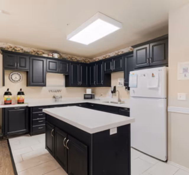 A modern kitchen with black cabinets and white countertops. The kitchen features a central island, a white refrigerator with papers attached, a microwave, a sink, and two large beverage dispensers on the counter. The ceiling has a large rectangular light fixture, and there is floral wallpaper trim near the ceiling.