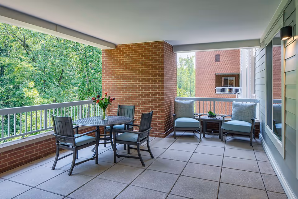 Covered outdoor patio area with a round table and four chairs on the left side, and two cushioned armchairs with a small table between them on the right side. The patio has a brick column and railing, overlooking green trees and another brick building in the background.